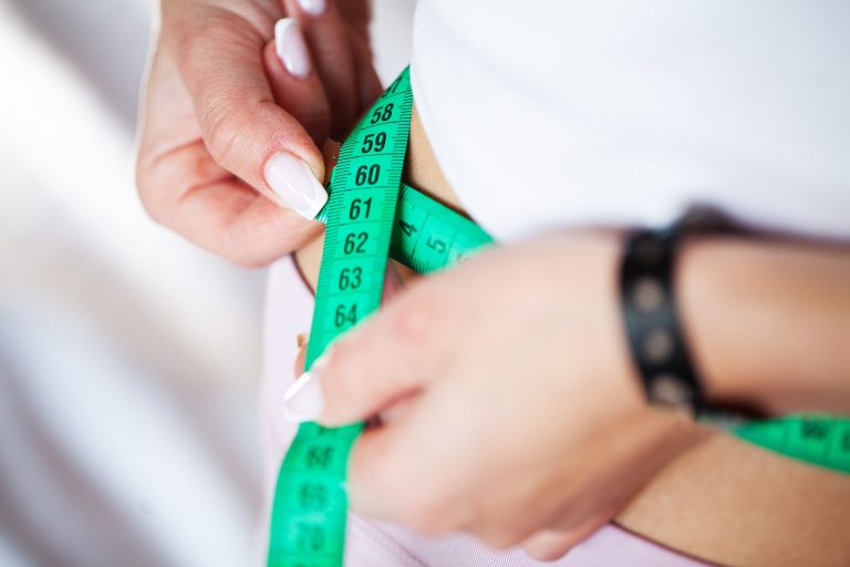 Slim young woman measuring her thin waist with a tape measure at home